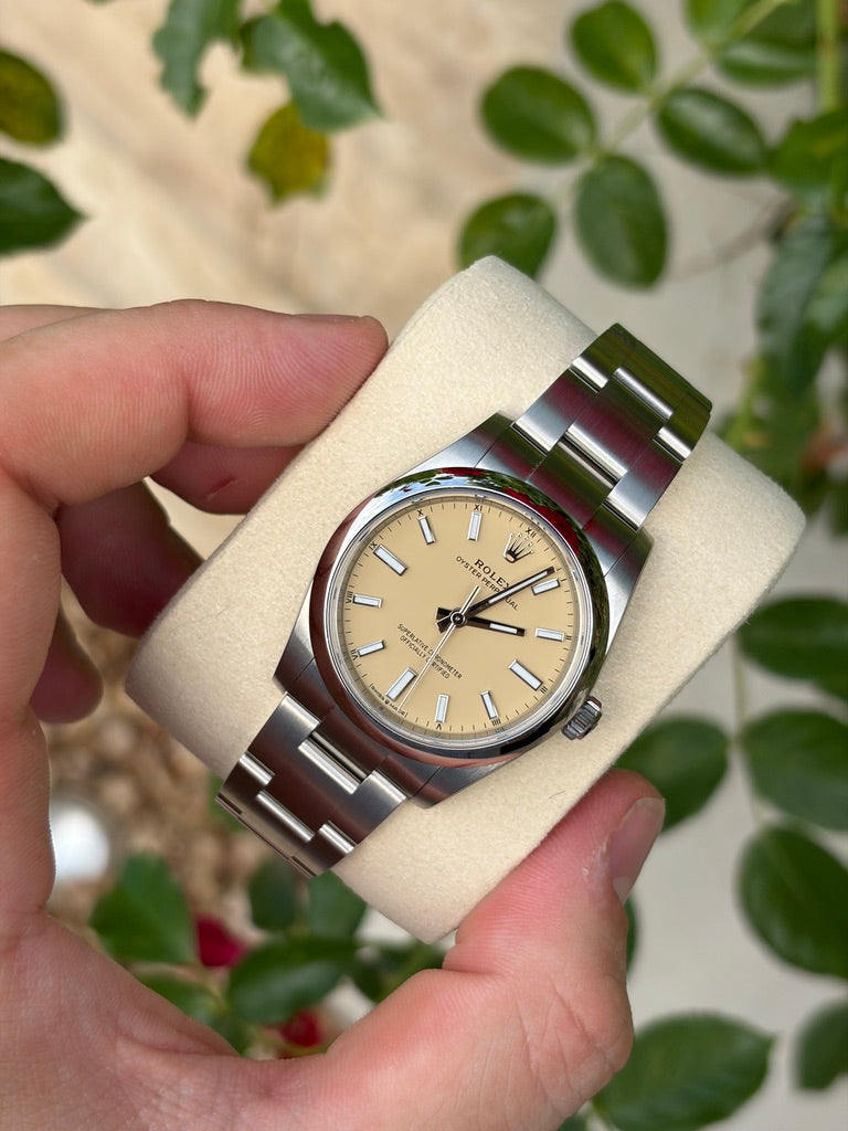 Rolex Oyster Perpetual 34 mm, Referenz 124200, aus Edelstahl mit beigem Zifferblatt, glatter Lünette und Oyster-Armband, auf einem Uhrenkissen gehalten.