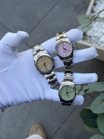 Drei Rolex Oyster Perpetual aus Edelstahl auf einem weißen Handschuh: 34 mm mit beigem Zifferblatt sowie zwei 31-mm-Modelle in Candy Pink und Pistachio.